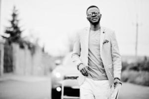 homme afro-américain riche et élégant en blazer et pantalon blanc, lunettes avec magazine à portée de main posé contre une voiture suv. photo