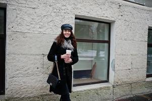 fille mexicaine bouclée en casquette en cuir et tasse de café en plastique à portée de main marchant dans les rues de la ville. photo