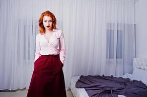 portrait d'une belle fille rousse en chemisier et jupe debout près du lit. photo