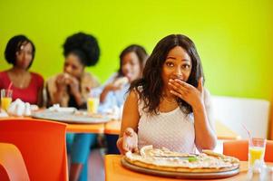femme africaine avec pizza assise au restaurant contre des filles à la peau foncée. visage surpris. photo