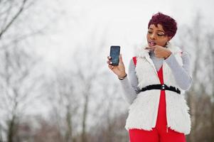 femme afro-américaine en pantalon rouge et veste de manteau de fourrure blanche posée le jour de l'hiver sur fond neigeux, montrer le doigt au téléphone. photo