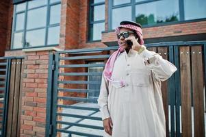 homme d'affaires arabe du moyen-orient posé dans la rue contre un bâtiment moderne avec des lunettes de soleil, parlant au téléphone mobile. photo
