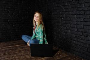 fille blonde élégante en veste et jeans avec ordinateur portable contre un mur noir en brique au studio. photo
