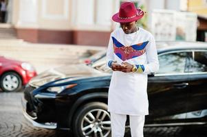 homme afro france élégant et branché en chapeau rouge et tenue blanche posée au jour de l'automne. homme d'affaires modèle noir africain avec téléphone portable à mains contre voiture de luxe.