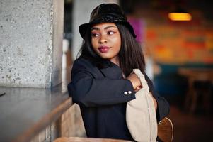 charmant modèle de femme afro-américaine en veste noire, chapeau et sac de taille relaxant au café pendant le temps libre. photo