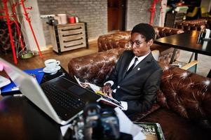 homme d'affaires afro-américain portant un costume noir et des lunettes assis au bureau avec un ordinateur portable et travaillant, lisant un magazine. photo