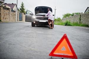 une jeune femme africaine triste se tient debout avec un triangle d'urgence contre une voiture avec un capot sauté. transport, problèmes de véhicules et concept de pannes. photo