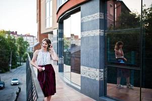 fille modèle blonde à la mode et belle dans une élégante jupe en velours rouge, chemisier blanc, posée avec téléphone et écouteurs contre les fenêtres du bâtiment de la ville. photo