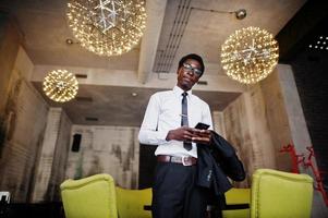 homme d'affaires afro-américain portant un costume noir et des lunettes au bureau regardant sur un téléphone mobile. photo