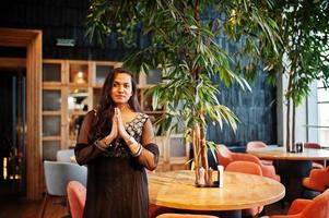 jolie fille indienne en robe sari noire posée au restaurant. montrant le signe des mains de namaste. photo