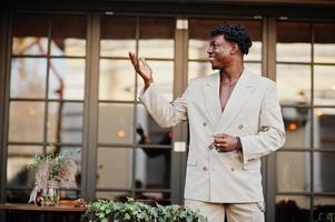 homme afro élégant en costume beige old school debout sur la terrasse du café. jeune homme africain à la mode en veste décontractée sur torse nu. photo