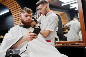 jeune homme barbu se coupe les cheveux par un coiffeur assis sur une chaise au salon de coiffure. âme de barbier. photo