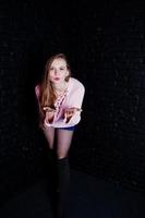 prise de vue en studio d'une fille brune au pull rose avec un short en jean contre un mur de briques noires. photo