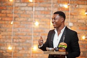 un jeune homme afro-américain respectable en costume noir tient un plateau avec un double hamburger et montre le pouce contre le mur de briques du restaurant avec des lumières. photo