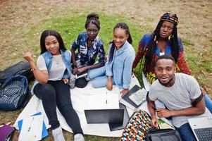 groupe de cinq étudiants africains qui passent du temps ensemble sur le campus de la cour universitaire. amis afro noirs assis sur l'herbe et étudiant avec des ordinateurs portables. photo