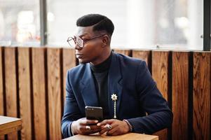 un homme afro-américain à la recherche étonnante porte un blazer bleu avec une broche, un col roulé noir et des lunettes posées dans la rue. gars noir à la mode assis derrière une table avec un téléphone portable. photo