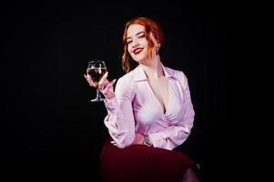 magnifique fille aux cheveux rouges en chemisier rose et jupe rouge avec un verre de vin à portée de main isolé sur noir. photo