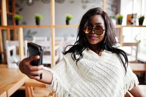 élégante fille afro-américaine à lunettes de soleil posée au café moderne et faisant selfie au téléphone.