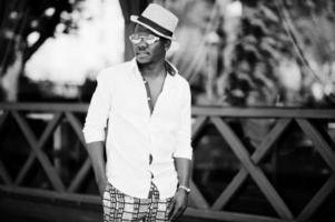 homme afro-américain élégant en chemise blanche et pantalon coloré avec chapeau et lunettes posés à l'extérieur. garçon modèle à la mode noir. photo