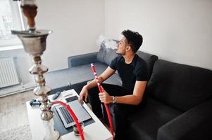 homme indien beau et à la mode en noir assis dans la chambre, fumant du narguilé, travaillant sur un ordinateur portable. photo