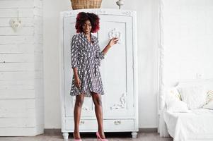 femme modèle afro-américaine haute à la mode avec des cheveux afro rouges en robe posée dans une salle blanche contre une armoire vintage. photo