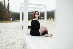 portrait de jeune fille brune en robe noire assis à la construction en bois blanc. photo