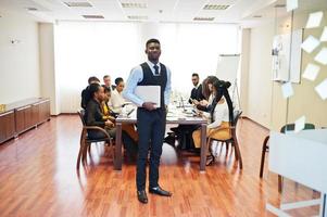 visage d'un bel homme d'affaires africain, tenant un ordinateur portable sur fond de réunion d'équipe multiraciale de gens d'affaires, assis dans une table de bureau. photo