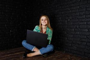 fille blonde élégante en veste et jeans avec ordinateur portable contre un mur noir en brique au studio. photo