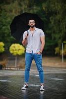 grand homme à la barbe arabe à la mode portant une chemise, un jean et des lunettes de soleil avec un parapluie posé à la pluie sur la place du parc. photo