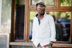 homme afro-américain élégant en chemise blanche et pantalon coloré avec chapeau et lunettes posés à l'extérieur. garçon modèle à la mode noir. photo