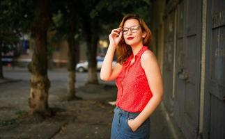 jolie femme rousse à lunettes, porter sur un chemisier rouge et une jupe en jean posant. photo