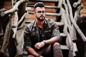 impressionnant beau grand homme macho barbe arabe à lunettes et veste militaire posé en plein air contre des escaliers en bois. photo