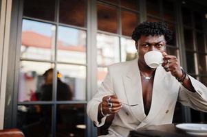 homme afro élégant en costume beige old school assis sur un café avec une tasse de café. jeune homme africain à la mode en veste décontractée sur torse nu. photo