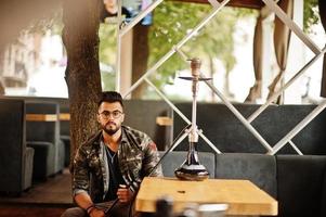 homme arabe à barbe élégante dans des verres et une veste militaire fumant le narguilé au bar de la rue. modèle arabe se reposant. photo