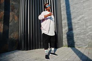 homme arabe hipster élégant à lunettes de soleil posé en plein air dans la rue au coucher du soleil contre un mur gris. photo