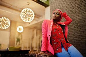 femme africaine élégante en chemise rouge, veste et chapeau posé café intérieur. photo