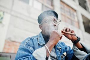 portrait en gros plan d'un homme afro-américain en veste de jeans, béret et lunettes, fumant un cigare. photo