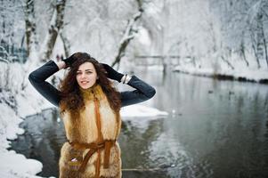 élégance fille bouclée en manteau de fourrure au parc forestier enneigé contre la rivière gelée en hiver. photo