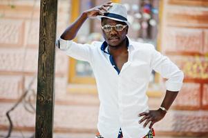 homme afro-américain élégant en chemise blanche et pantalon coloré avec chapeau et lunettes posés à l'extérieur. garçon modèle à la mode noir. photo