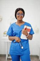 portrait d'une jeune femme médecin pédiatre afro-américaine heureuse en manteau uniforme bleu et stéthoscope avec des livres à portée de main. soins de santé, médecine, spécialiste de la médecine - concept. photo