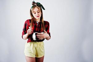 jeune femme au foyer drôle en chemise à carreaux et short jaune pin up style avec casserole et cuillère de cuisine isolé sur fond blanc. photo