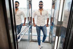 élégant grand modèle d'homme arabe en chemise blanche, jeans et lunettes de soleil posés à l'intérieur de l'ascenseur. photo