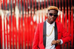 modèle d'homme afro-américain de mode en costume rouge, avec des cheveux en surbrillance et un sac à main posé dans la rue et regardant ses montres. photo