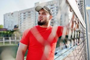 jeune homme musclé barbu brutal portant une chemise rouge, un short et une casquette au stade. photo