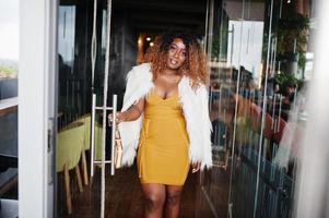 femme afro-américaine glamour en robe jaune et cape en laine blanche avec sac à main posé à l'entrée de la porte vitrée du restaurant. photo