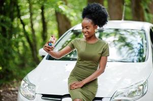 femme afro-américaine posée contre une voiture blanche sur une route forestière et regardant un téléphone portable. photo