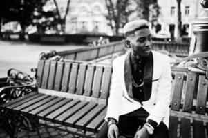 chic bel homme afro-américain en costume blanc assis sur un banc. photo