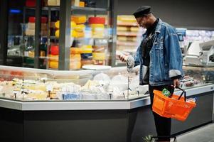 élégant homme afro-américain décontracté à la veste en jean et au béret noir tenant un panier, debout près du réfrigérateur à fromage et faisant ses courses au supermarché. photo