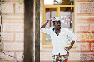 homme afro-américain élégant en chemise blanche et pantalon coloré avec chapeau et lunettes posés à l'extérieur. garçon modèle à la mode noir. photo