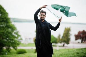 l'homme pakistano bangladesh porte des vêtements traditionnels noirs et des lunettes pose à l'extérieur, tenant le drapeau du pakistan. photo